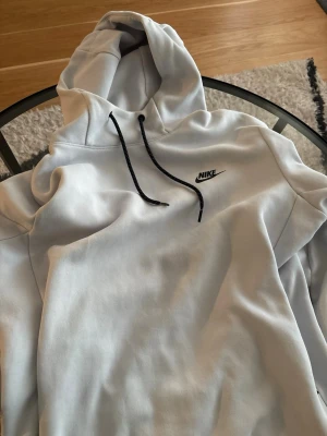 Vit hoodie från Nike med logga - Snygg vit hoodie från Nike med svart logga på bröstet och svarta dragsnören i huvan. Klassisk passform och mjukt material som känns skönt mot huden. Perfekt för dig som gillar sportig och clean stil.