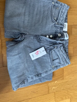 Grå raka jeans från 157 - Snygga grå jeans från 157 med rak passform och hög midja. Jeansen har klassisk femficksdesign, knäppning med flera silverfärgade knappar och är tillverkade i denim. Perfekta för en trendig och avslappnad look.