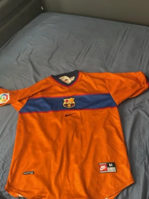 Barcelona matchtröja orange Nike - Snygg orange FC Barcelona matchtröja från Nike med blått och rött band över bröstet och klubbmärke i mitten. Tröjan har korta ärmar, V-ringad krage med blå och röda detaljer och är tillverkad i ett lätt funktionsmaterial.