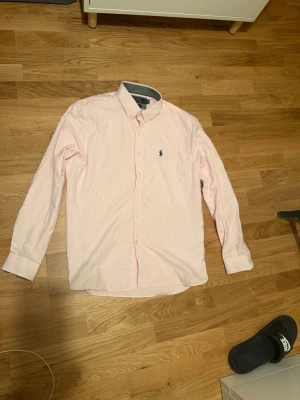 Ljusrosa skjorta från Ralph Lauren - Klassisk ljusrosa skjorta från Ralph Lauren med lång ärm och knappar framtill. Skjortan har en broderad blå logga på bröstet och är tillverkad i bomull. Perfekt för en clean och stilren look.