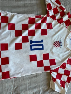 Kroatien fotbollströja Modrić  - Säljer en vit och röd fotbollströja från Nike med Kroatiens landslagsmärke på bröstet och Modrić samt nummer 10 i blått på ryggen. Tröjan har klassiskt rutmönster i rött och vitt och är tillverkad i lätt, andasvänligt material. Perfekt för match eller träning. Köpt på marknad i Kroatien för 30 euro