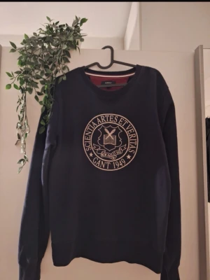 Mörkblå sweatshirt från Gant - Mörkblå sweatshirt från Gant med rund halsringning och stort tryckt emblem på bröstet med texten 'Scientia Artes Et Veritas Gant 1949'. Tröjan har långa ärmar och ribbade muddar vid ärmslut och nederkant. Perfekt för en avslappnad och stilren look.