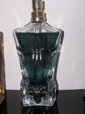Jean Paul Gaultier Le Male parfym - En ikonisk parfymflaska formad som en manlig torso i klart glas med mörkgrön vätska och guldfärgad spraytopp. Flaskan har en unik och konstnärlig design som verkligen sticker ut i badrumshyllan.