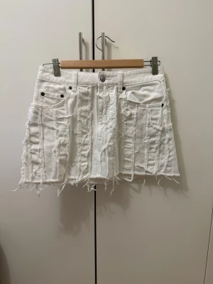 Vit kort jeanskjol med fransar - Säljer en vit kort jeanskjol med råa kanter och fransiga detaljer. Kjolen har flera fickor fram och bak samt dekorativa sömmar och slitningar för en edgy look. Perfekt för dig som gillar streetstyle och vill sticka ut.