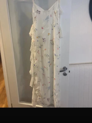 Vit långklänning med volanger och blommor - Romantisk vit långklänning med tunna axelband och volangdetaljer längs hela sidan. Klänningen har ett blommigt mönster i pastellfärger och är tillverkad i ett lätt, luftigt material som passar perfekt till sommaren.