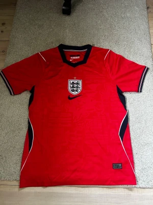 England Vm tröja 2026 - Snygg röd England matchtröja från Nike med svart och vit detalj vid kragen och ärmsluten. Tröjan har England-emblemet på bröstet och är tillverkad i lätt och ventilerande Aero-Fit material, perfekt för fotboll eller träning.
