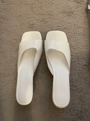 Vita slip-in sandaler med fyrkantig tå - Stilrena vita slip-in sandaler med bred rem och fyrkantig tå. Sandalerna har en låg klack och är tillverkade i ett glansigt material som ger en modern look. Perfekta för dig som gillar minimalistisk och trendig stil.