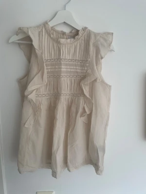 Beige ärmlös blus med volang - Jättesöt beige blus i tunn bomull med volangdetaljer längs sidorna och spetsinslag. Blusen är ärmlös och har rundad hals med små veck framtill. Perfekt för en romantisk och luftig stil. Den är sparsamt använd och har inga hål🥰