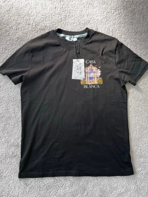 Svart t-shirt från Casablanca - Snygg svart t-shirt från Casablanca med tryck framtill som visar ett klassiskt valv med blommor och hav i bakgrunden. T-shirten har rund hals och korta ärmar. Perfekt för dig som vill ha en stilren men unik look.