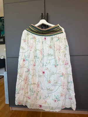 Vit långkjol med broderade blommor - Säljer en vit långkjol med färgglada broderade blommor och ett bredare, virkat midjeband i grönt, rosa och blått. Kjolen har en luftig och flowy känsla, perfekt för varma dagar. Passar dig som gillar boho-stil och unika detaljer.
