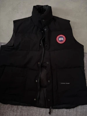 Svart dunväst från Canada Goose - Snygg svart dunväst från Canada Goose med hög krage och klassisk logotyp på bröstet. Västen har två stora fickor framtill och stängs med både dragkedja och tryckknappar. Perfekt för lager-på-lager och håller dig varm under kyliga dagar.