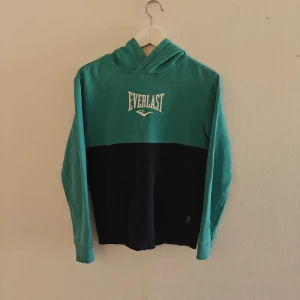 Everlast hoodie - Snygg hoodie från Everlast i turkos och mörkblått med vit logga på bröstet. Tröjan har huva, lång ärm och en stor magficka framtill. Tillverkad i 100% bomull, perfekt för chill dagar eller träning.
