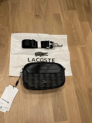 Svart axelväska från Lacoste - Snygg svart axelväska från Lacoste med geometriskt mönster och silverfärgade detaljer. Väskan har justerbar axelrem i textil och dragkedja upptill. Tillverkad i slitstarkt material och har en svart Lacoste-logga framtill. Perfekt för att bära det viktigaste.