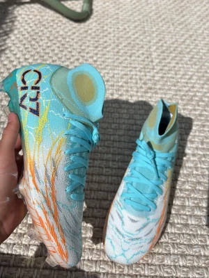 Nike CR7 Mercurial fotbollsskor - Snygga Nike Mercurial CR7 fotbollsskor i ljusblått, vitt och gult med högt skaft och coola grafiska detaljer. Skorna har CR7-logga på sidan och genomskinlig sula med färgstänk och Nike-logga undertill. Perfekta för dig som vill sticka ut på planen.
