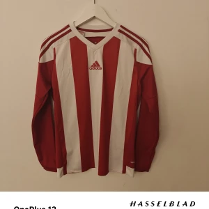 Adidas fotbollströja - Snygg långärmad fotbollströja från Adidas i rött och vitt med klassiska vertikala ränder och v-ringad krage. Tillverkad i lätt och ventilerande ClimaCool-material, perfekt för intensiva matcher eller träning. Ikoniska Adidas-ränder på axlar och ärmar.