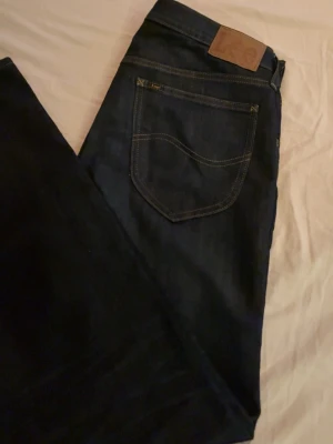 Mörkblå jeans från Lee - Säljer ett par mörkblå jeans från Lee med klassisk femficksdesign och snygga kontrastsömmar. Jeansen har en rak passform och är tillverkade i slitstarkt denimtyg. Perfekta för dig som gillar tidlös och enkel stil. Knappt använda, är som nya!!!