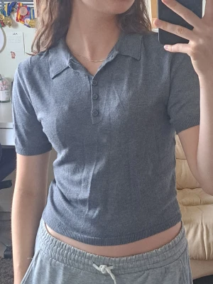 Grå polotopp från H&M Divided - Säljer en grå polotopp från H&M Divided i storlek S. Toppen har korta ärmar, krage och knappar framtill. Materialet är mjukt och stretchigt, perfekt till jeans eller kjol. Enkel och stilren design som passar till många olika looks.