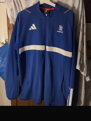 Blå Adidas France ziptröja - Säljer en blå ziptröja från Adidas med France-logga på bröstet och vita detaljer över bröstet och ärmarna. Tröjan har hög krage, dragkedja framtill och två sidofickor. Perfekt för sport eller chill. Materialet är polyester.