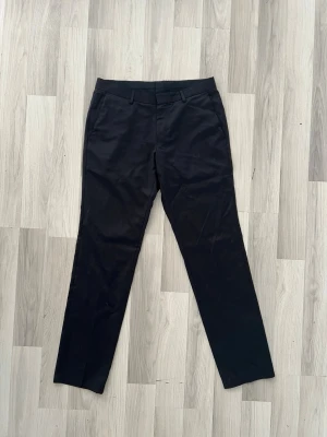 Svarta raka kostymbyxor Hugo Boss - Snygga svarta kostymbyxor med raka ben och klassisk passform. Byxorna har hällor för skärp, dragkedja och metallknapp i midjan. Perfekta för en stilren look. Materialet känns slätt och dressat. Fråga mig om storlek