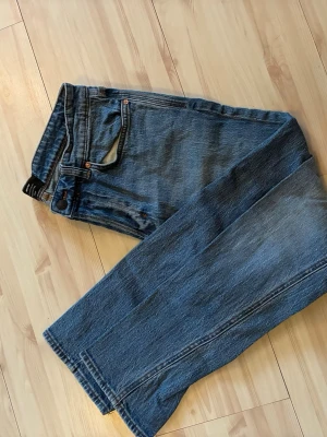 Blå raka jeans med hög midja - Klassiska blå jeans med rak passform och hög midja. Jeansen har fem fickor, bälteshällor och stängs med knappar. Tillverkade i slitstark denim som ger en tidlös look. Perfekta för dig som gillar en enkel och clean stil.