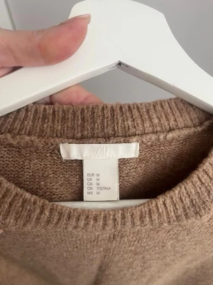Beige stickad tröja från H&M - Mysig beige stickad tröja från H&M i storlek M. Tröjan har rund halsringning, långa ärmar och ribbade muddar vid ärmslut och nederkant. Perfekt för lager på lager och enkel att matcha med jeans eller kjol.