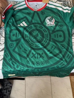 Mexiko landslagströja Adidas grön - Säljer en grön Mexiko landslagströja från Adidas med aztekiskt mönster och vita detaljer. Tröjan har rund hals med röd kant, vita ränder på axlarna och Mexikos fotbollsförbunds logga på bröstet. Perfekt för dig som älskar fotboll och vill sticka ut på planen.