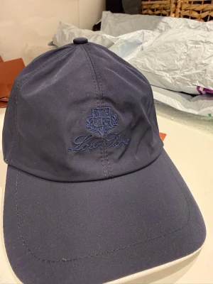 Mörkblå keps från Loro Piana - Snygg mörkblå keps från Loro Piana med broderad logga framtill och justerbar rem bak. Klassisk böjd skärm och stilren design. Tillverkad i ett mjukt, slitstarkt material som känns lyxigt och passar perfekt till streetwear eller casual outfits.