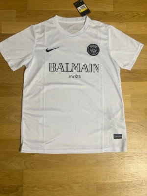 Vit PSG x Balmain t-shirt Nike - Snygg vit t-shirt från samarbetet mellan PSG och Balmain, med svart Nike-logga och PSG-emblem på bröstet. T-shirten har klassisk rund hals och korta ärmar. Materialet är mjukt och lätt polyester, perfekt för en sportig look.