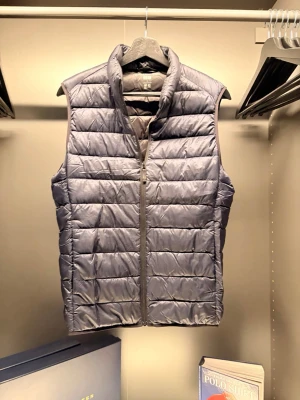 Uniqlo - Snygg grå dunväst med quiltad design och hög krage. Västen har en rak passform och stängs med dragkedja framtill. Perfekt att slänga över en hoodie eller tröja för extra värme. Materialet är lätt och smidigt, vilket gör den enkel att bära och styla.