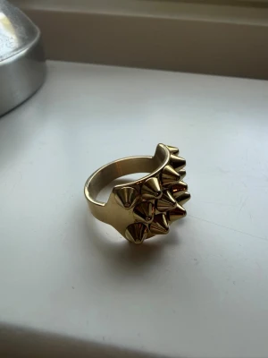 Helt ny Edblad ring  - En helt ny Edblad ring . Den är aldrig använd. Cool ring i guldfärgad metall med flera små konformade nitar på ovansidan. Ringen har en chunky design och ger ett edgy intryck. Perfekt för dig som vill sticka ut med dina accessoarer.
