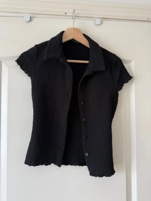Svart kortärmad skjorta Gina Tricot - Svart skjorta från Gina Tricot i storlek XS. Skjortan har korta ärmar, krage och knäppning framtill. Materialet är strukturerat och stretchigt vilket ger en cool look. Perfekt att styla med jeans eller kjol.