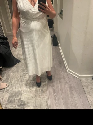 Vit omlottklänning med v-ringning - Elegant vit omlottklänning med djup v-ringning fram och bak. Klänningen har ett fint fall, markerad midja och är ärmlös. Materialet ser ut att vara glansigt och lätt, troligen polyester eller satin. Perfekt för sommarens tillfällen.