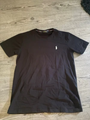 Svart t-shirt från Polo Ralph Lauren - Klassisk svart t-shirt från Polo Ralph Lauren med den ikoniska vita logon broderad på bröstet. T-shirten har rund halsringning och korta ärmar. Tillverkad i mjuk bomull för en bekväm känsla och passar perfekt till alla tillfällen. Pris kan diskuteras 