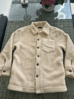 Beige teddyjacka från GANT - Supermjuk teddyjacka från GANT i beige färg med klassisk skjortkrage, bröstficka och knäppning framtill. Jackan har långa ärmar med knappar vid ärmslut och är tillverkad i fluffigt teddy-material och är perfekt för kallare sommarkvällar eller vår/höst. Den är knappt använd. 
