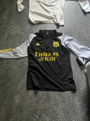 Olympique Lyonnais Adidas - Svart träningsjacka från Adidas med Olympique Lyonnais-logga på bröstet. Jackan har vita ärmar med gula detaljer och dragkedja vid halsen. Tillverkad i funktionsmaterial som passar perfekt till träning. Adidas och Emirates Fly Better-tryck på framsidan. Olympique Lyonnais Adidas