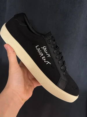 Svarta Saint Laurent sneakers 44 - Svarta sneakers från Saint Laurent med vit broderad logga på sidan och vit sula. Skorna har en klassisk låg profil, svarta skosnören och detaljer i både canvas och läder. Perfekta för dig som gillar stilrena och exklusiva sneakers.