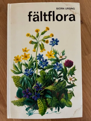 Fältflora av Björn Ursing - Fältflora av Björn Ursing är en praktisk fälthandbok med färgillustrationer av svenska växter. Omslaget är vitt med en färgglad teckning av olika blommor. Perfekt för dig som vill lära dig mer om växter i naturen.