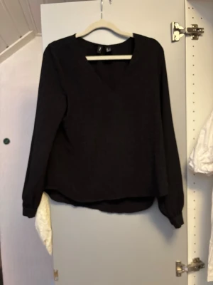 Svart blus med v-ringning från Vero Moda - Stilren svart blus från Vero Moda med v-ringad hals och långa ärmar. Blusen har en enkel, rak passform och är tillverkad i ett lätt, vävt material som ger en snygg siluett. Perfekt att matcha med jeans eller kjol för en trendig look.
