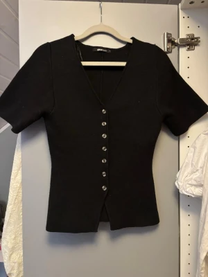 Svart stickad  kortärmad tröja Gina Tricot - Stilren svart tröja från Gina Tricot med korta ärmar och djup V-ringning. Koftan har dekorativa knappar framtill och är figurnära i modellen. Perfekt att matcha med jeans eller kjol för en trendig look. Använd max 5 gånger 
