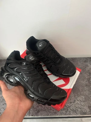 Nike Air Max Plus helsvarta sneakers - Säljer ett par helsvarta Nike Air Max Plus sneakers med ikoniska vågiga detaljer och synliga Air-enheter i sulan. Skorna har mesh- och syntetmaterial i ovandelen, svarta snören och små silvriga Swoosh-loggor på sidorna. Perfekta för dig som gillar streetwear och sportig stil.