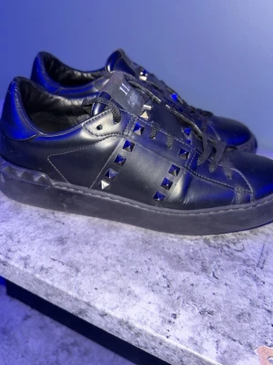 Svarta Valentino Rockstud sneakers äkta - Säljer ett par svarta Valentino Rockstud sneakers i läder med coola nitar längs sidorna. Skorna har svarta skosnören och en chunky sula som ger en edgy vibe. Perfekta för dig som vill sticka ut med en lyxig och unik look.