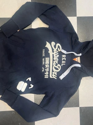 Mörkblå Superdry vintage hoodie - Snygg mörkblå hoodie från Superdry med stort vitt tryck på bröstet och japansk text. Hoodien har en klassisk huva med gråa dragsnören och en känguruficka framtill. Perfekt för en avslappnad och trendig stil. Tröjan är rätt så liten i storleken 