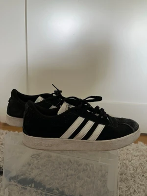 Svarta Adidas sneakers med vita ränder - Klassiska svarta Adidas sneakers med de ikoniska tre vita ränderna på sidan. Skorna har lågt skaft och är gjorda i mockaliknande material med vit sula. Perfekta för en avslappnad och sportig stil.