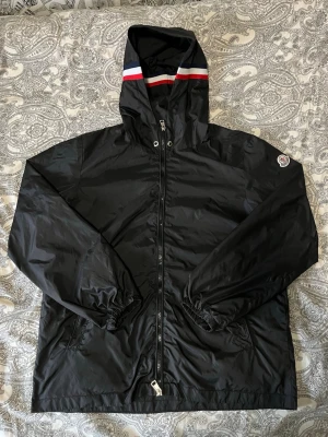 Svart vindjacka från Moncler - Snygg svart vindjacka från Moncler med huva och dragkedja framtill. Jackan har Moncler-logga på ärmen och en cool detalj med röd och vit rand på huvan. Tillverkad i lätt, glansigt material som står emot vind. Perfekt för dig som vill ha en stilren och sportig look.