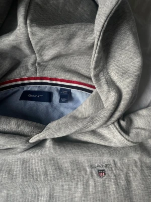 Grå hoodie från GANT  - Snygg grå hoodie från GANT med broderad logga och sköld på bröstet. Klassisk modell med huva, känguruficka och ribbade muddar. Tillverkad i mjuk bomullsblandning för en bekväm känsla. Perfekt för en avslappnad och stilren look.