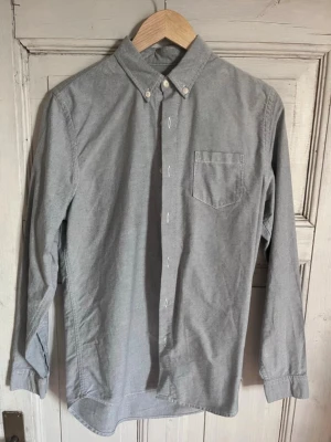 Grå skjorta från Folk med knappar - Säljer en stilren grå skjorta från Folk med klassisk button-down krage och bröstficka. Skjortan har långa ärmar och vita knappar framtill. Perfekt för dig som gillar enkel och clean design. Materialet är mjukt och känns som bomull.