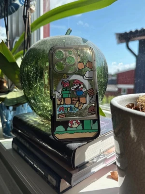 Genomskinligt Mario mobilskal (Iphone 16 pro) - Säljer ett genomskinligt mobilskal med färgglatt Super Mario-motiv. Skalet har illustrationer av Mario, svampar och klassiska block från spelet. Passar till en mobil med tre kameralinser. Tillverkat i tålig plast och har en cool retro gaming vibe.