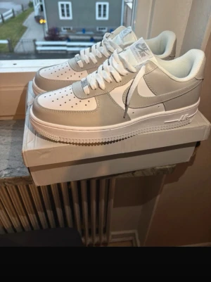Nike Air Force 1 vita/grå skor - Nike Air Force 1 i vitt och ljusgrått med klassisk siluett. Skorna har perforerad tå, vita skosnören och den ikoniska Swoosh-loggan på sidan. Ovandelen är i läder och textil, med vadderad krage för extra komfort. Perfekta för dig som gillar sportig och stilren look.