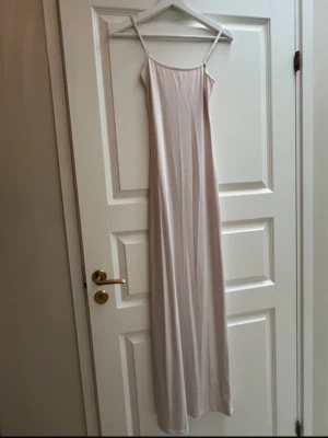 Ljus långklänning med tunna band - Säljer en elegant ljus långklänning med tunna axelband och rak passform. Klänningen har minimalistisk design och är tillverkad i ett mjukt, slätt material som faller fint längs kroppen. Perfekt för sommarens alla tillfällen.