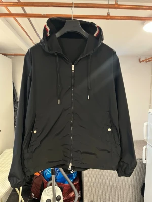 Svart vindjacka från Moncler - Snygg svart vindjacka från Moncler med huva och dragkedja framtill. Jackan har två fickor med tryckknappar och justerbar dragsko i huvan. Detaljer med Monclers signaturrand på insidan av huvan. Perfekt för blåsiga dagar. Har två ytterst små skador på båda fickor därav pris sänkningen !